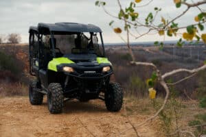 A black and lime green Honda utility-terrain vehicle (UTV) with Carlstar Versa Trail tires parked on a dirt trail in a rural or wilderness area. The overcast sky adds a muted light, and sparse vegetation surrounds the UTV. The scene conveys a sense of isolation and remoteness.