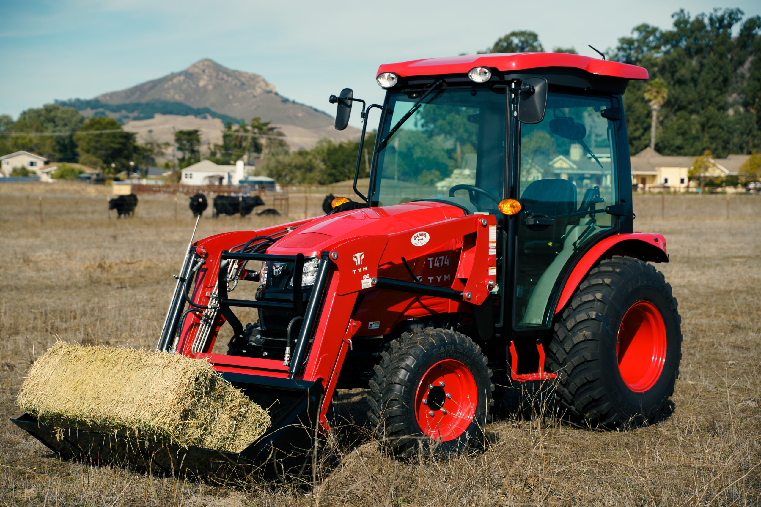 A red TYM tractor with a front loader carrying a bale of hay on Carlstar Versa Turf Ag tires, positioned in a field with cows and houses in the background, under a clear sky with a mountain visible at a distance.